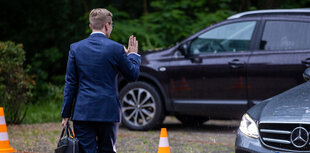 ein Mann mit Aktentasche und erhobener rechter Hand vor zwei Autos