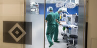 Blick durch eine Glastür mit HSV-Log in einen Operationssal mit technischen Geräten und zwei Menschen in grüner Kleidung