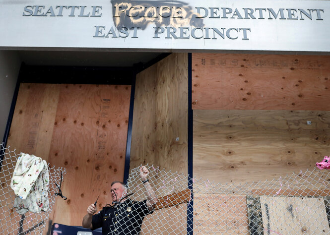 Ein Mann entfernt einen Zaun vor einem Haus, das mit Holz verbarrikadiert ist. Das Haus ist eine Polizeistation in Seattle das Wort "Police" wurde übersprüht mit "people"