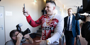 Rapper Gzuz mit Goldkette und Rolex filmt lachend einen Fotografen im Gericht