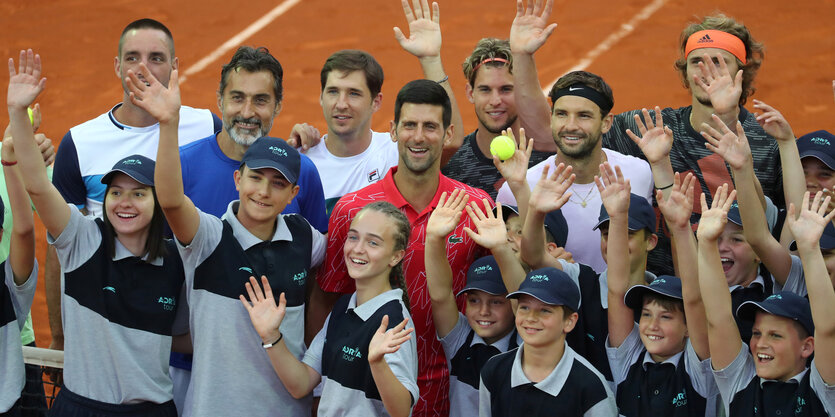 Tennisspieler und Ballkinder winken eng beieinanderstehend in eine Kamera