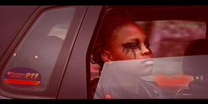Blick durch ein Autofenster auf das Gesicht einer jungen Frau, Augen schwarz umrandet.