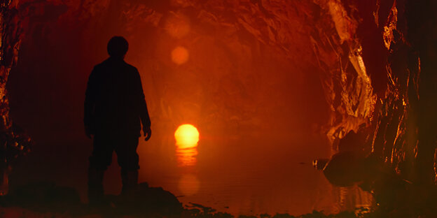 Ein Mann steht in einer Höhle mit unterirdischem See, in dem eine Sonne versinkt.