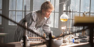 Rosamund Pike als Marie Curie beugt sich über einen Tisch mit verschiedenen Flüssigkeiten in Reagenzgläsern.