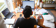 Ein Mensch sitzt am Schreibtisch im Home Office, vor zwei Laptops, einem Smartphone und Dokumentenstapeln.