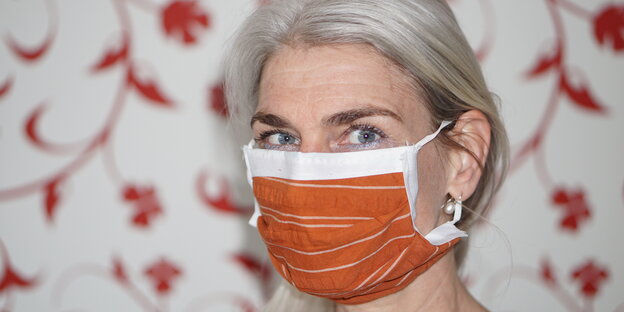 Petra Muth, eine Frau mit orangenem Mund-Nasen-Schutz. Im Hintergrund eine Tapete mit roten Ästen und Blättern. Petra Muth, eine Frau mit orangenem Mund-Nasen-Schutz. Im Hintergrund eine Tapete mit roten Ästen und Blättern.