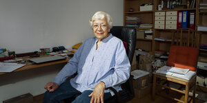 Eine ältere Frau sitzt auf einem Schreibtischstuhl und lächelt. Im Hintergrund ein Schreibtisch und ein Regal mit Ordnern.