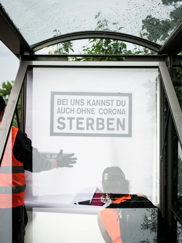 Zwei Menschen in Warnwesten entrollen ein Werbeplakat an einer geöffneten Vitrine: "Bei uns kannst du auch ohne Corona sterben", steht darauf Zwei Menschen in Warnwesten entrollen ein Werbeplakat an einer geöffneten Vitrine: "Bei uns kannst du auch ohne Corona sterben", steht darauf