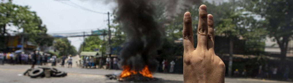 Demonstrierende Menschen in Myanmar tragen ein großes Transparent der regierungschefin. Die Menschen heben ihre Hände in die luft. Kleinerfinger und Daumen sind in die Handflächen geklappt, so dass nur Zeigefinger bir Ringfinger in die Luft stehen.