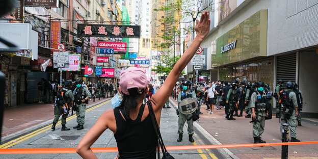Eine Frau stemmt die linke Hand in die Hüfte. Den rechte Arm streckt sie in die Luft. Vor ihr ist ein Absperrband. Dahinter Polizisten auf einer Straße.