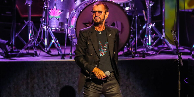 Ringo Starr als alter Mann. Er hat dunkles Haar und einen dunklen Bart. Er steht auf einer Bühne und hält ein Mikrofon in der Hand, hinter ihm steht ein Schlagzeug.