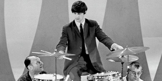 Ringo Starr als junger Mann. Er spielt gerade Schlagzeug. Neben ihm auf den Knien ist Ed Sullivan, der zu Starr aufblickt.