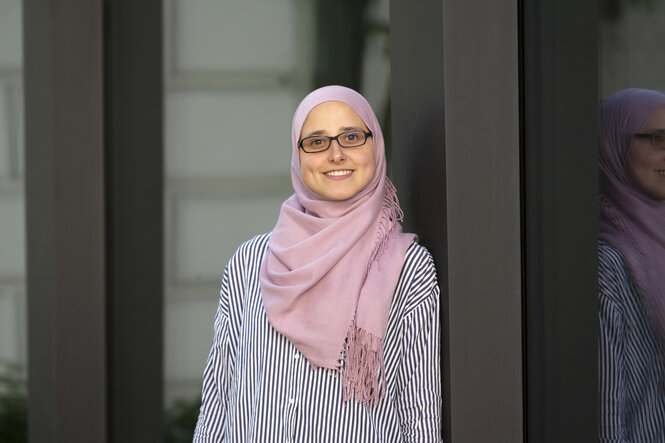 Fatima El Sayed, eine junge Frau, steht in einem Türrahmen. Sie lächelt und trägt eine Brille und ein Kopftuch.