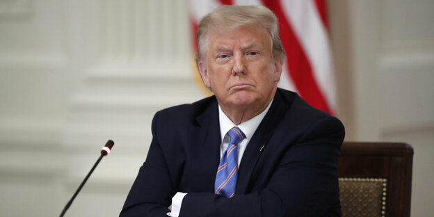 Donald Trump sitzt an einem Tisch, vor ihm ein Mikrofon, hinter ihm eine Fahne der USA. Er hat einen roten Kopf und kleine wässrige Augen. Seine Nase ist schmal. Er schaut missbilligend und gelangweilt. Seine Mundwinkel sind weit nach unten gezogen.