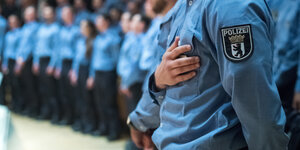 Viele uniformierte Polizist:innen stehen bei ihrer Vereidigung in einem Saal, einer hält sich seine Hand an die linke Brust