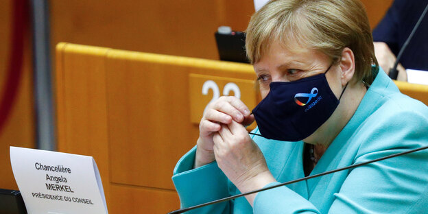 Angela Merkel rückt eine dunkelblaue Maske mit dem Logo der deutschen Ratspräsidentschaft zurecht