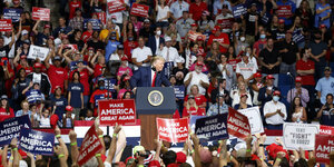 Trump auf einem Podium während einer Wahlkampfvranstaltung, im ihn herum viele Menschen mit Plakaten