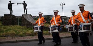 Männer in orangen Jacken und mit Trommeln marschieren entlang einer Statue in Derry