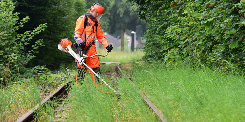 eine Mann in roter Schutzkleidung mäht das wild wachsende Gras zwischen Gleisen