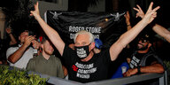 Roger Stone mit "Free Roger Stone"-NasenMundschutz und "Free Roger Stone"-T-Shirt hebt hinter einer Absperrung beide Arme in die Höhe und macht mit den Fingern Victory-Zeichen. Neben ihm stehen seine Anhänger