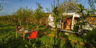 Auf einer Obstwiese stehen Campingstühle und ein Bus, ein Mädchen liest