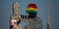 Aktivist:innen mit Kletterausrüstung sitzen auf einer hohen Skulptur aus Stahl. Die Figur tragt eine Sturmmaske in Regenbogenfarben