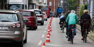 links sind Autofahrer von hinten zu sehen, rechts ein durch rote Hütchen abgesperrter Radweg, auf dem Radfahrer fahren