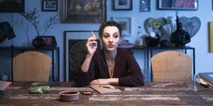 Kunstudentin Leila Raabe im Poträt: sie sitzt rauchend an einem Tisch, hinter ihr hängen viele Bilder