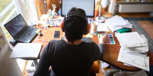 Eine Frau sitzt im Homeoffice vor ihrem Laptop an einem Schreibtisch voller Papiere. Sie ist von hinten abgebildet