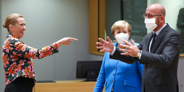 Eine Frau zeigt lachend auf einen Mann, der die Arme entgegenstreckt. Merkel mit Maske guckt zuz
