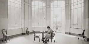 Schwarzweißfotografie: Frau schreibt Brief an einem Tische mitten in einem großen leeren Raum mit hohen Fenstern