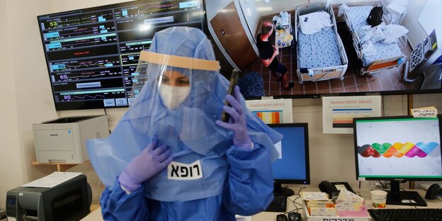 Frau mit Pexiglasgesichtsschutz, Maske, Ganzkörperschutz und Handy in einem Krankenhaus