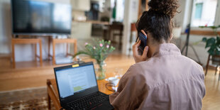 EIne Frau sitzt zuhause vor ihrem Computer und telefoniert mit dem Handy