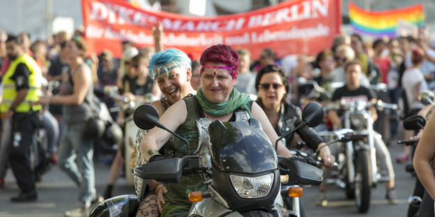Zwei Frauen sitzen auf einem Motorrad, umringt von Demonstrantinnen und Bannern und Regenbogenfahnen – ein Bild vom Dyke March für lesbische Sichtbarkeit