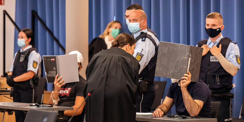 Zwei Angeklagte halten Mappen als Sichtschutz vor ihre Gesichter, während eine Verteidigerin sich mit dem rechten Angeklagten bespricht und Justizbeamte hinter den Angeklagten stehen