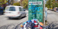 Ein Kleiderberg liegt vor einem Altkleidercontainer mit der Aufschrift „Kleiderbox“.