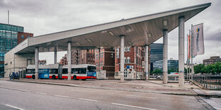 Spitz zulaufendes Dach auf Säulen, darunter ein Bus, dahinter Backsteingebäude, davor eine Straße