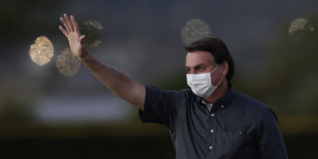 Jair Bolsonaro hebt die Hand und winkt, er trägt einen weißen Mundschutz-