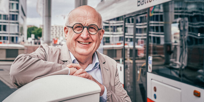 Ein Mann mit Glatze stützt sich mit dem Unterarm auf, hinter ihm ein Bus