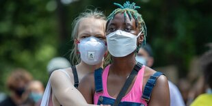 Zwei Frauen mit Maske umarmen sich. Eine hat eine dunkle, die andere eine helle Hautfarbe.