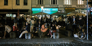 viele Menschen sitzen und stehen vor einer nächtlichen Häuserfassade auf der Straße, im Hintergrund eine Leuchtschrift "Napoli"