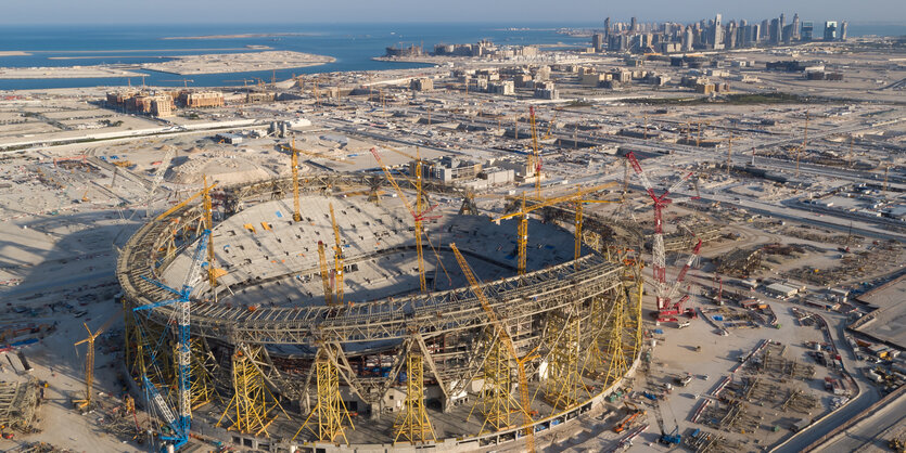 Luftaufnahme einer Stadionbaustelle, weiter hinten sind das Meer und schicke Hochhäuser