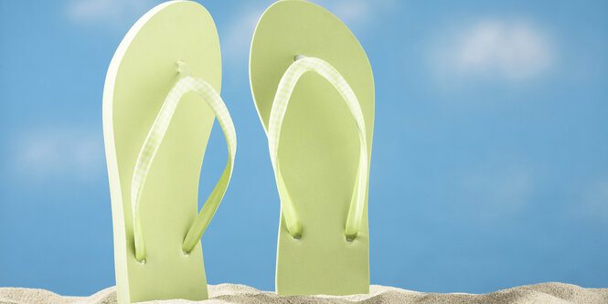 Ein ppar weisse FlipFlops stecken im Sand vor blauem Himmel