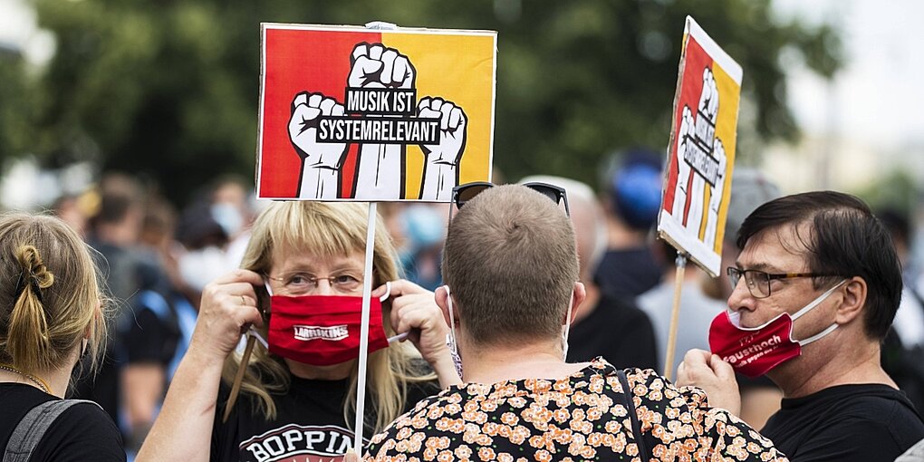 Menschen mit roten Mundschutzmasken stehen in einer menschenmenge und halten Transparente mit der Aufschrift "Musik ist systemrelevant"