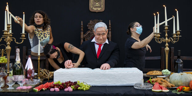 eine Figur, die wie Netanjahu aussiseht, hinter einer reich gedeckten Tafel, an der Frauen Kerzen anzünden