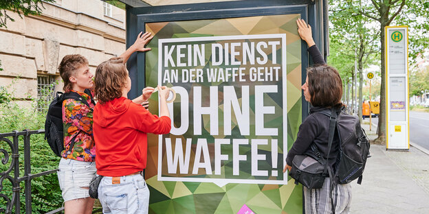 Aktivist:innen kleben ein Plakat auf eine Bushaltestelle. Darauf steht: "Kein Dienst an der Waffe geht ohne Waffe"