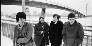 Die vier Musiker der Band Joy Division stehen vor einer Betonbrücke, ganz rechts Ian Curtis