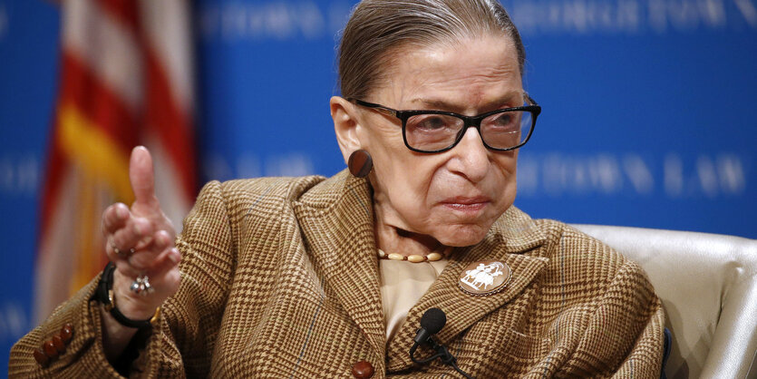 Portrait von Ruth Bader Ginsburg, die lange Haare hat und zu einem Zopf gebunden hat, dazu trägt sie ein Cord-Sakko und eine Brille