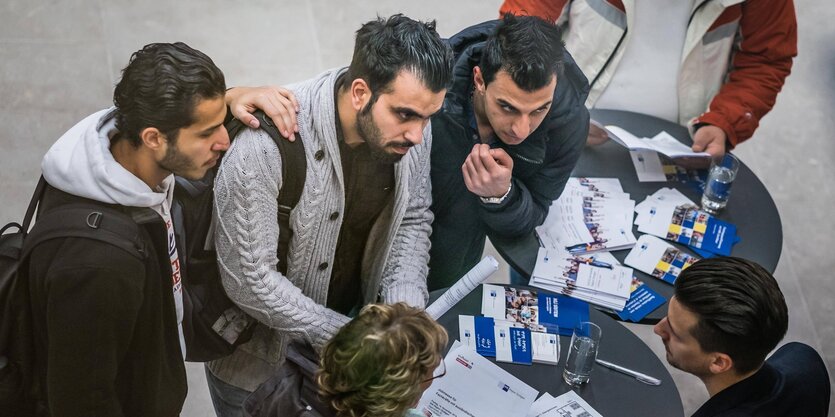 Männer stehen um einen Tisch und hören einem anderen Mann in Anzug zu