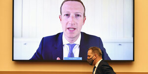 Das Konterfei von Mark Zuckerbberg ist auf einem riesigen Bildschirm zu sehen, an dem ein Mann mit Maske vorbeiläuft